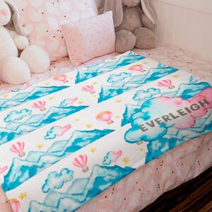May include: A white fleece blanket with a watercolor design of blue mountains, pink clouds, and hot air balloons. The blanket has a pink cloud with the name "EVERLEIGH" written in brown text.