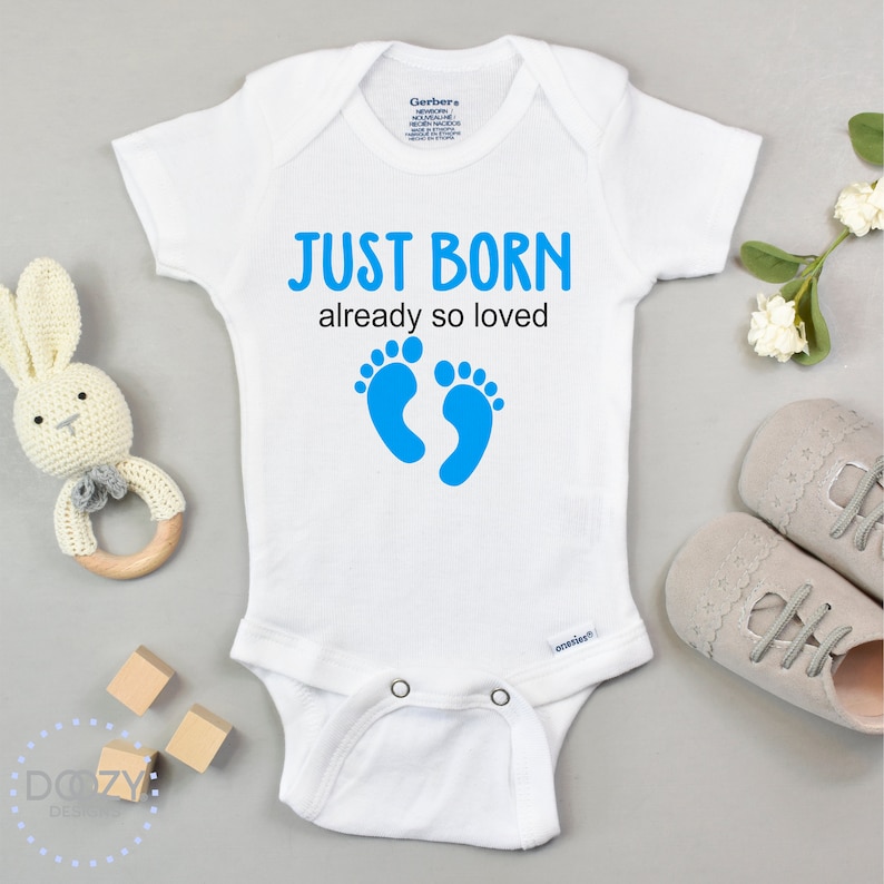 May include: White baby onesie with short sleeves and blue footprints. The onesie has the text "JUST BORN already so loved" printed on it.