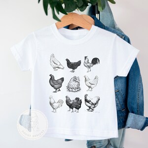 Pode incluir: Camiseta branca com uma ilustra&ccedil;&atilde;o em preto e branco de diferentes galinhas. O desenho inclui um galo, galinhas e pintinhos.