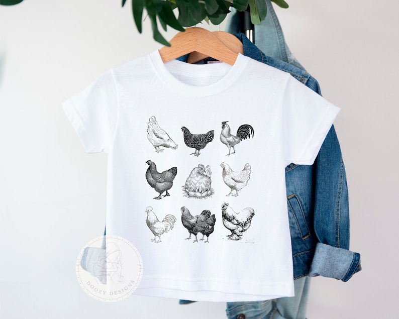 Pode incluir: Camiseta branca com uma ilustra&ccedil;&atilde;o em preto e branco de diferentes galinhas. O desenho inclui um galo, galinhas e pintinhos.