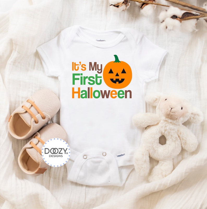 Puede incluir: Un body blanco para beb&eacute; con el texto "It's My First Halloween" en verde, naranja y marr&oacute;n. Una cara de calabaza naranja sonriente est&aacute; al lado del texto.