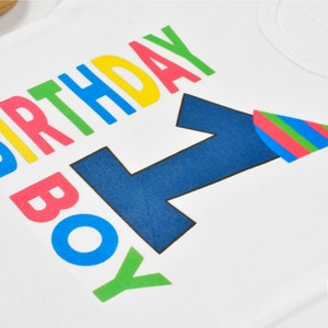 May include: White onesie with the words "Birthday Boy" in rainbow colors and a blue number one with a party hat on top.