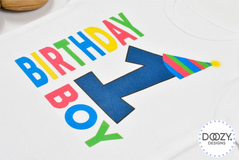 May include: White onesie with the words "Birthday Boy" in rainbow colors and a blue number one with a party hat on top.