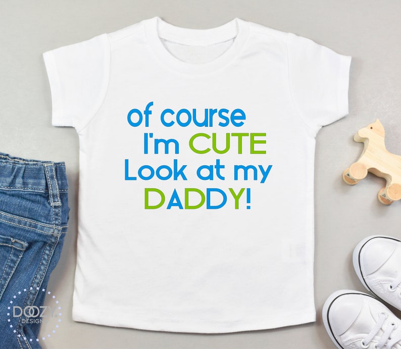 May include: White baby t-shirt with blue and green text that reads "of course I'm CUTE Look at my DADDY!"