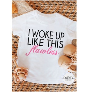 May include: White toddler t-shirt with the text "I WOKE UP LIKE THIS flawless" in black and pink. The shirt is displayed with a pair of tan baby shoes and a wooden toy on a woven surface. The Doozy Designs logo is visible.