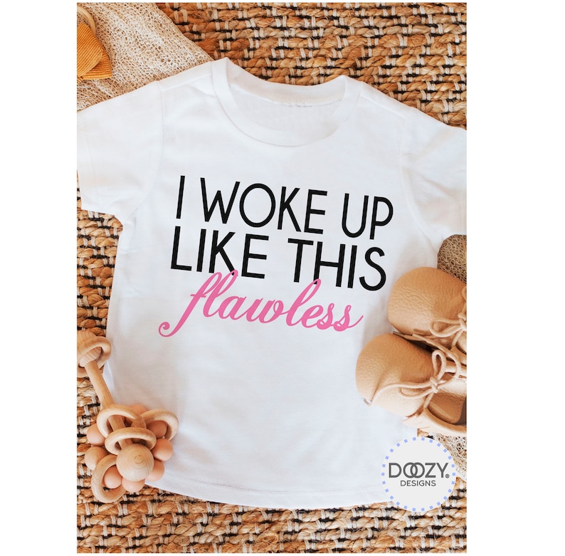 May include: White toddler t-shirt with the text "I WOKE UP LIKE THIS flawless" in black and pink. The shirt is displayed with a pair of tan baby shoes and a wooden toy on a woven surface. The Doozy Designs logo is visible.