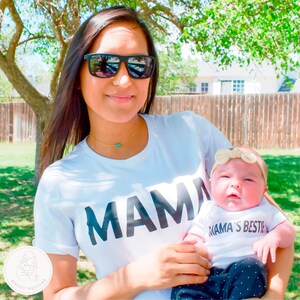 May include: A woman wearing a white t-shirt with the word "MAMA" printed on it is holding a baby wearing a white t-shirt with the text "MAMA'S BESTIE" printed on it. The baby is wearing a beige headband with a bow.