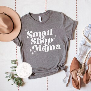 K&ouml;nnte beinhalten: Ein graues T-Shirt mit dem Text "Small Shop Mama" in Wei&szlig;, mit einem wei&szlig;en Pluszeichen auf jeder Seite der W&ouml;rter. Das Shirt liegt flach auf einer wei&szlig;en Oberfl&auml;che mit einem braunen Hut, einem Paar braunen Schuhen und einer wei&szlig;en Halskette.