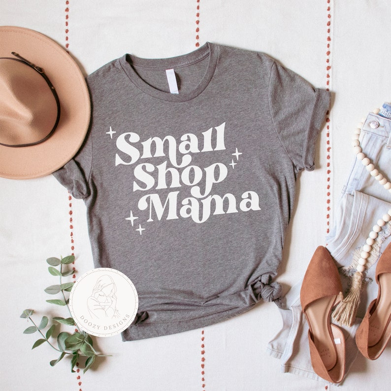 K&ouml;nnte beinhalten: Ein graues T-Shirt mit dem Text "Small Shop Mama" in Wei&szlig;, mit einem wei&szlig;en Pluszeichen auf jeder Seite der W&ouml;rter. Das Shirt liegt flach auf einer wei&szlig;en Oberfl&auml;che mit einem braunen Hut, einem Paar braunen Schuhen und einer wei&szlig;en Halskette.