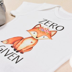Op de afbeelding: Witte baby romper met een vos die een bloemenkrans draagt en de tekst "ZERO GIVEN".