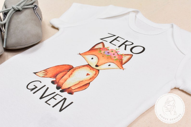 Op de afbeelding: Witte baby romper met een vos die een bloemenkrans draagt en de tekst "ZERO GIVEN".