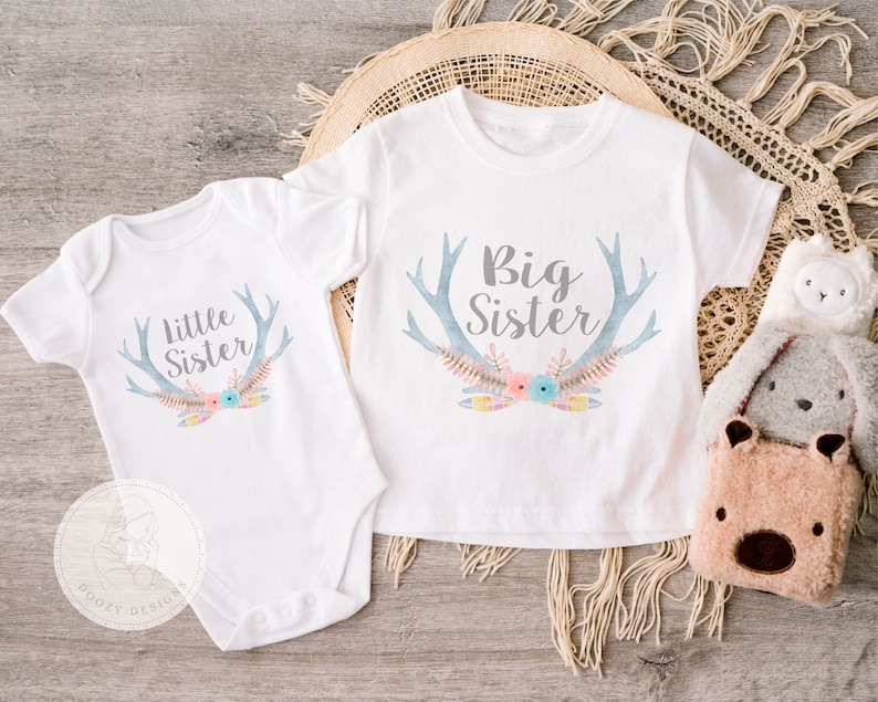 May include: Two white shirts with a blue and gray antler design and floral accents. One shirt says "Big Sister" and the other says "Little Sister".