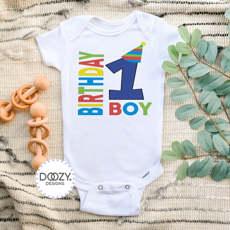 May include: White baby onesie with a colorful "Birthday Boy" graphic featuring a blue number one with a party hat. The onesie has a white background and is decorated with a colorful birthday graphic.