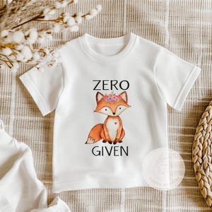 Op de afbeelding: Wit T-shirt met een cartoonvos die een bloemenkrans draagt. De tekst "ZERO GIVEN" is onder de vos gedrukt.