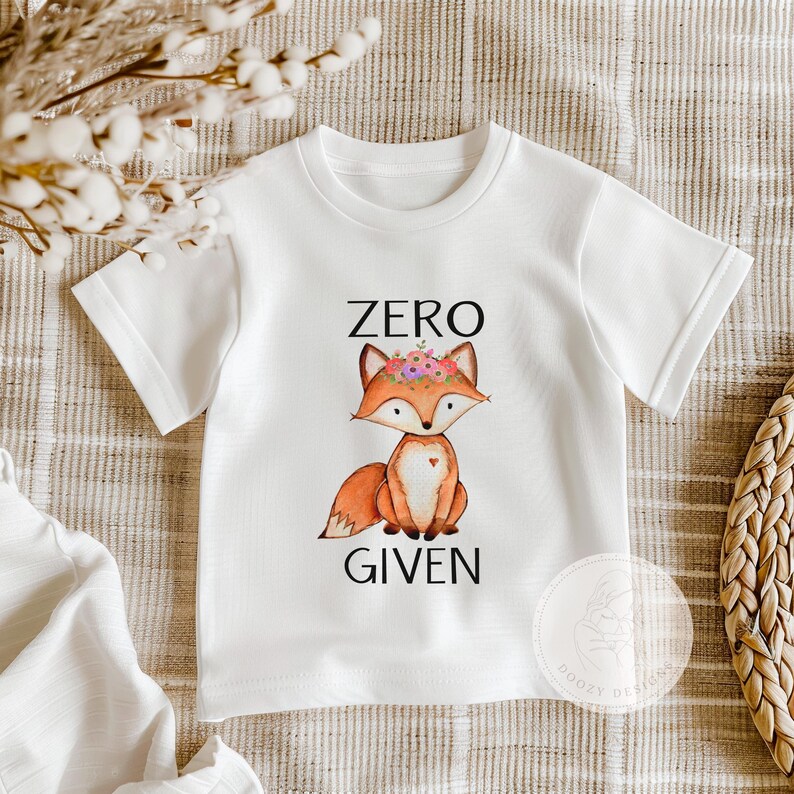 Op de afbeelding: Wit T-shirt met een cartoonvos die een bloemenkrans draagt. De tekst "ZERO GIVEN" is onder de vos gedrukt.