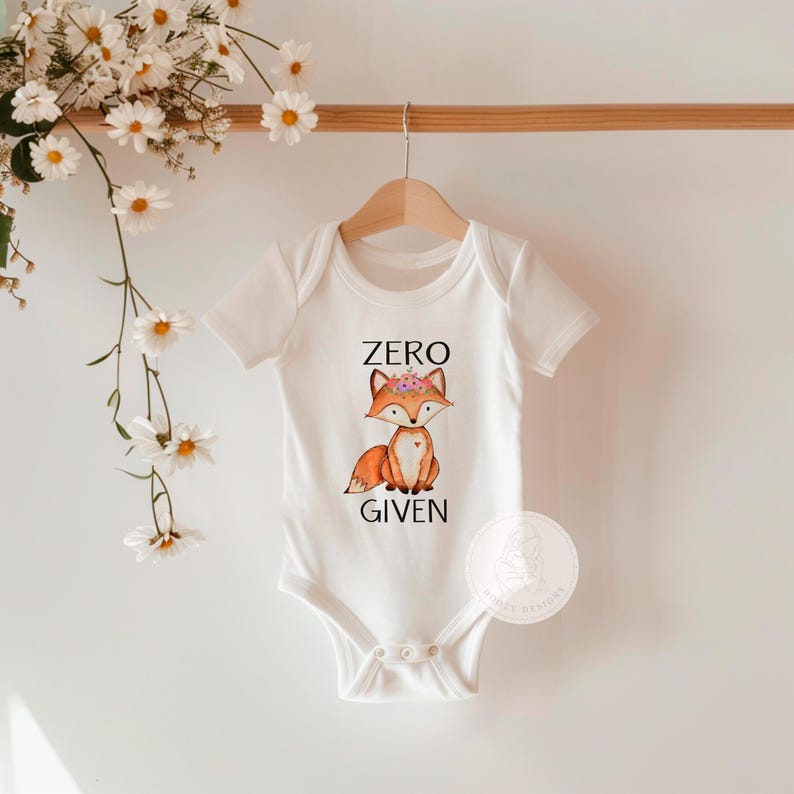 Op de afbeelding: Witte baby romper met een vos die een bloemenkrans draagt en de tekst "ZERO GIVEN".