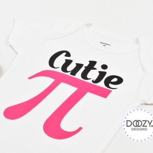 Pu&ograve; includere: Un body bianco per neonato con un simbolo pi rosa e la parola "cutie" in testo nero. Il body ha un piccolo logo nell'angolo in basso a destra che dice "Doozy Designs".
