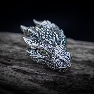 Puede incluir: Un colgante de cabeza de dragón de plata con ojos verdes. El colgante está detallado con escamas y tiene una superficie texturizada.