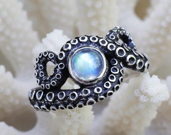 May include: A silver-toned ring with an octopus design, featuring a central, round, iridescent stone. The ring's tentacles wrap around the stone. The ring is set against a white, textured background, resembling coral.