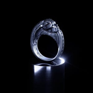May include: A silver-toned ring featuring a sculpted human face integrated into the band. The metal surface displays intricate, textured detailing along the sides and a polished finish, capturing light against a dark, solid background.