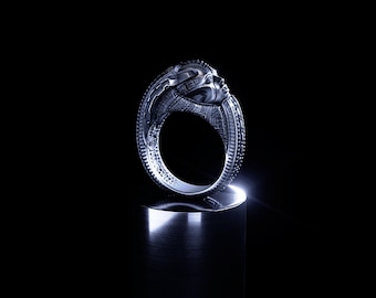 May include: A silver-toned ring featuring a sculpted human face integrated into the band. The metal surface displays intricate, textured detailing along the sides and a polished finish, capturing light against a dark, solid background.
