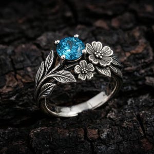 May include: A silver ring featuring a round, bright blue gemstone set among sculpted metal leaves and two small flowers. The band is detailed with textured botanical elements that wrap around the finger, creating a nature-inspired design.
