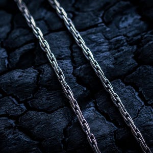 May include: Two silver chains lay side by side on a dark, textured surface. The chains are made of small, linked metal pieces.