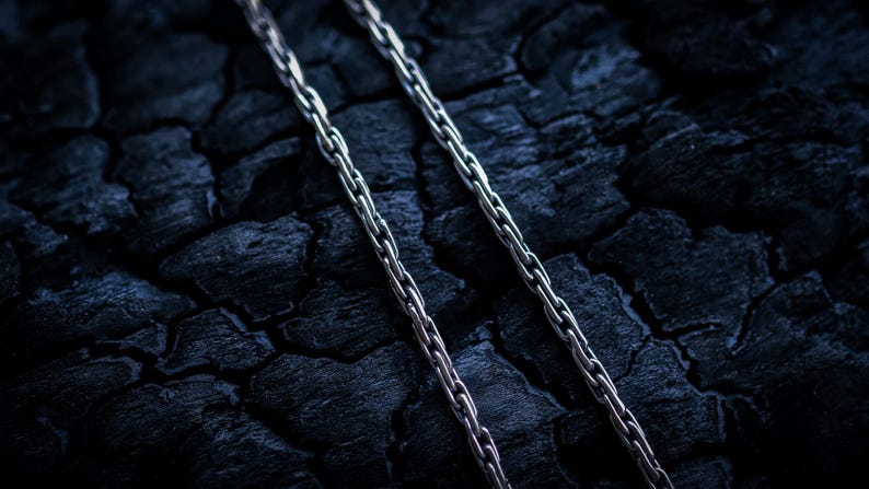 May include: Two silver chains lay side by side on a dark, textured surface. The chains are made of small, linked metal pieces.