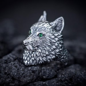 Puede incluir: Un anillo de tono plateado que presenta una cabeza de lobo esculpida y detallada con patrones de pelaje texturizados y dos pequeños ojos de piedra preciosa de color verde vibrante. El metal tiene un acabado oxidado que resalta los rasgos.