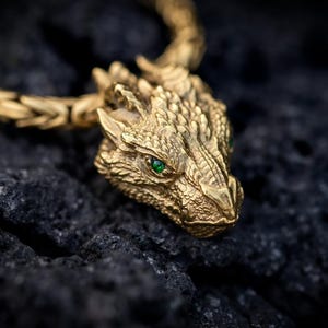 May include: A gold-toned dragon head pendant attached to a thick, woven Byzantine chain. The dragon features intricate scale detailing and small, vibrant green gemstone eyes, creating a highly textured and detailed metallic appearance.