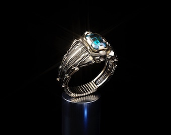 May include: A metallic ring with an intricate, textured band featuring a rectangular, faceted blue gemstone set at the top. The ring displays a complex, industrial-style design with deep grooves and raised details, catching light with a bright glint.