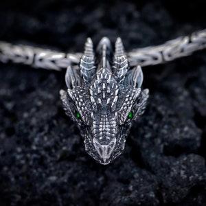 May include: A silver dragon pendant with green eyes, detailed with scales and spikes, hangs from a silver chain.