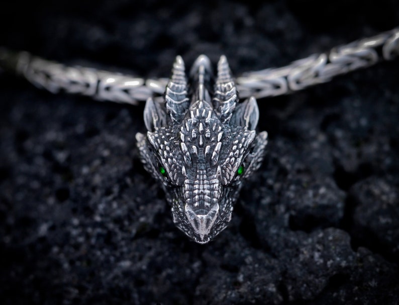May include: A silver dragon pendant with green eyes, detailed with scales and spikes, hangs from a silver chain.