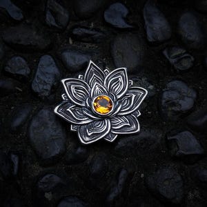 May include: A silver-toned metal brooch shaped like a multi-layered lotus flower with intricate engraved detailing on each petal. A single, round, faceted amber-colored gemstone is set securely in the center of the floral design.