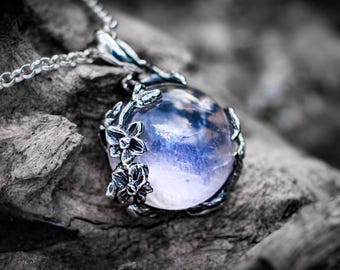 Pode incluir: Um colar com pingente de prata apresentando uma pedra preciosa redonda, translúcida e de cor púrpura pálida, engastada em uma moldura de metal escuro decorada com detalhes florais esculpidos. O pingente pende de uma delicada corrente de prata.