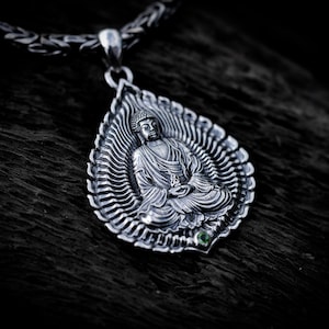 Op de afbeelding: Een zilveren hanger met een zittende Boeddha-figuur in een meditatieve houding, gecentreerd in een bladvormig frame met stralende lijnen. De hanger bevat een kleine groene edelsteen aan de basis en hangt aan een dikke, gevlochten zilveren ketting.