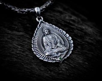 May include: A silver pendant featuring a seated Buddha figure in a meditative pose, centered within a leaf-shaped frame with radiating lines. The pendant includes a small green gemstone at the base and hangs from a thick, woven silver chain.
