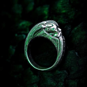 May include: A silver ring featuring a detailed, elongated creature head with a smooth, curved cranium and intricate, ribbed textures along the band. The metallic surface reflects a vibrant green light, highlighting the sculpted, biomechanical features.