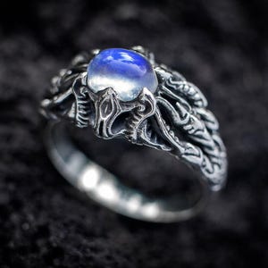 Könnte beinhalten: Ein Silberring mit einem zentralen, runden, irisierenden blauen Mondstein-Cabochon. Die Ringschiene und die Fassung sind mit aufwendigen, texturierten Metallarbeiten versehen, die an organische, verzweigte Formen oder Tentakel erinnern.