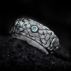 May include: A silver ring featuring an intricate, raised woven pattern across the entire band. A single, small, round, vibrant green gemstone is set securely in the center of the metalwork, contrasting with the dark, oxidized silver finish.