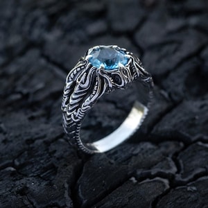 Puede incluir: Un anillo de plata con una piedra preciosa redonda y facetada de color azul claro engastada en una banda de metal texturizada y detallada. La banda muestra intrincados patrones de relieve orgánicos y fluidos alrededor del engaste.