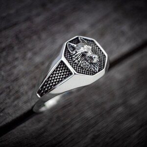 Puede incluir: Un anillo de tono plateado con una cara octogonal que presenta un relieve detallado de una cabeza de lobo. El fondo de la cabeza de lobo tiene una textura de patrón de rayado cruzado, y los lados del anillo también tienen un diseño a juego.
