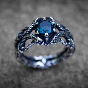 Könnte beinhalten: Ein silberfarbener Ring mit einem dunkelblauen, rund geschliffenen Edelstein in der Mitte. Die Ringschiene ist mit einem detaillierten, strukturierten Design versehen, das an zwei ineinander verschlungene Schlangen erinnert, deren Köpfe den Stein halten.