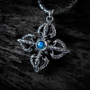 May include: A silver pendant featuring a four-pronged vajra design with intricate metalwork and a central round iridescent blue stone. The pendant is suspended from a thick, woven silver chain, showcasing detailed craftsmanship and a polished finish.