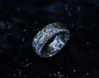 May include: A silver-toned metal ring featuring a complex, raised geometric pattern across its entire outer surface. The design consists of interconnected rectangular lines and recessed grooves, creating a textured, grid-like appearance on the band.