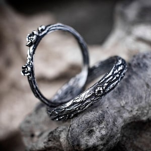 May include: Two silver-toned rings featuring a textured, bark-like surface finish. One ring stands upright, displaying small, raised floral details along its band, while the second ring lies flat, showcasing a continuous, rugged, branch-like pattern.