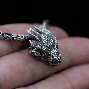 May include: A silver dragon pendant necklace with green eyes. The dragon is detailed with scales and has a textured surface. The necklace is made of a silver chain.