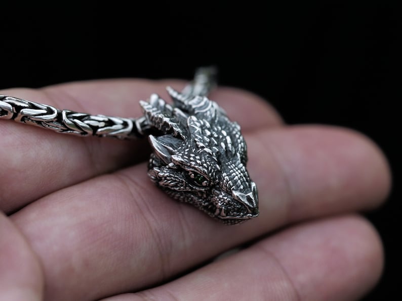 May include: A silver dragon pendant necklace with green eyes. The dragon is detailed with scales and has a textured surface. The necklace is made of a silver chain.
