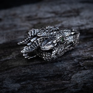May include: A silver dragon head ring with a green gemstone eye. The ring is detailed with scales and teeth.
