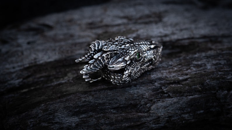 May include: A silver dragon head ring with a green gemstone eye. The ring is detailed with scales and teeth.